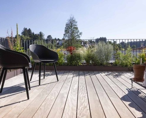 Holzterrasse im Dachgarten Holzterrasse im Dachgarten mit modernen schwarzen Gartenstühlen, bepflanzten Beeten und Blick auf die umliegenden Häuser.